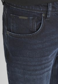 Vaqueros de denim azul oscuro con una textura suave, que cuentan con un bolsillo frontal y una pequeña etiqueta de marca. Patrones de decoloración sutiles visibles.