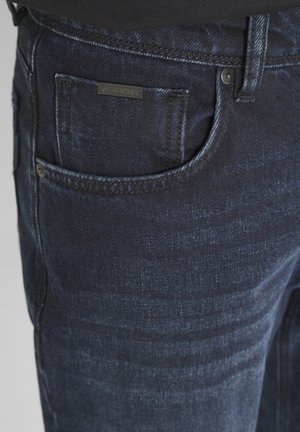 Vaqueros de denim azul oscuro con una textura suave, que cuentan con un bolsillo frontal y una pequeña etiqueta de marca. Patrones de decoloración sutiles visibles.