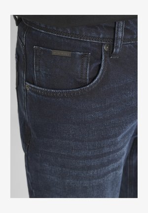 Tamno plave traperice s glatkom teksturom, s prednjim džepom i malom markiranom oznakom. Vidljivi su suptilni uzorci istrošenosti.