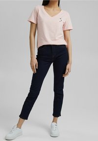 Camiseta de cuello en V de color rosa claro con mangas cortas, que presenta un gráfico de un pájaro negro en el pecho. Combinada con jeans ajustados azul oscuro y zapatillas blancas.