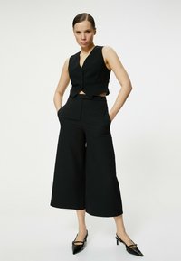 Zwarte op maat gemaakte vest met een cropped ontwerp en knopen aan de voorkant, gecombineerd met wijde zwarte broeken. Puntige zwarte hakken maken de look af.
