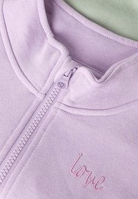 Gros plan d'un vêtement lavande avec une fermeture éclair, un col doux et le mot « love » brodé avec un fil assorti.