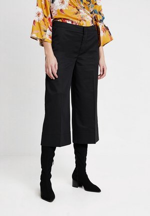 Nainen pukeutuneena mustiin leveälahkeisiin, lyhennettyihin housuihin, mustiin korkokenkiin ja keltaiseen kukkakuosiseen puseroon, seisomassa valkoista taustaa vasten.