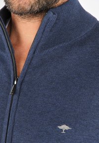 Sudadera azul marino con cremallera y cuello alto, fabricada en un tejido de punto texturizado. Presenta un discreto logo de árbol en el pecho.