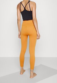 Mujer de pie descalza sobre una esterilla de yoga, vistiendo un top deportivo negro con tirantes y leggings naranjas de tiro alto, vista desde atrás.