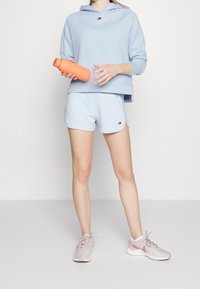 Jasnoniebieska bluza z kapturem i logo, dopasowane szorty i jasnoróżowe sneakersy. Trzyma pomarańczową butelkę na wodę. Gładka faktura materiału.