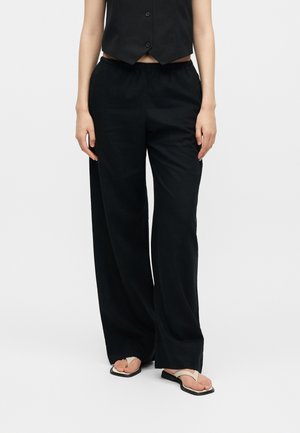 Γυναίκα με φαρδιά μαύρη παντελόνα, μαύρο γιλέκο με κουμπιά και μπεζ ανοιχτόχρωμα σανδάλια σε απλό λευκό φόντο.