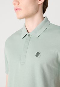Polo-shirt vert clair avec un col texturé et une patte de boutonnage. Présente un logo circulaire avec une lettre stylisée en noir.