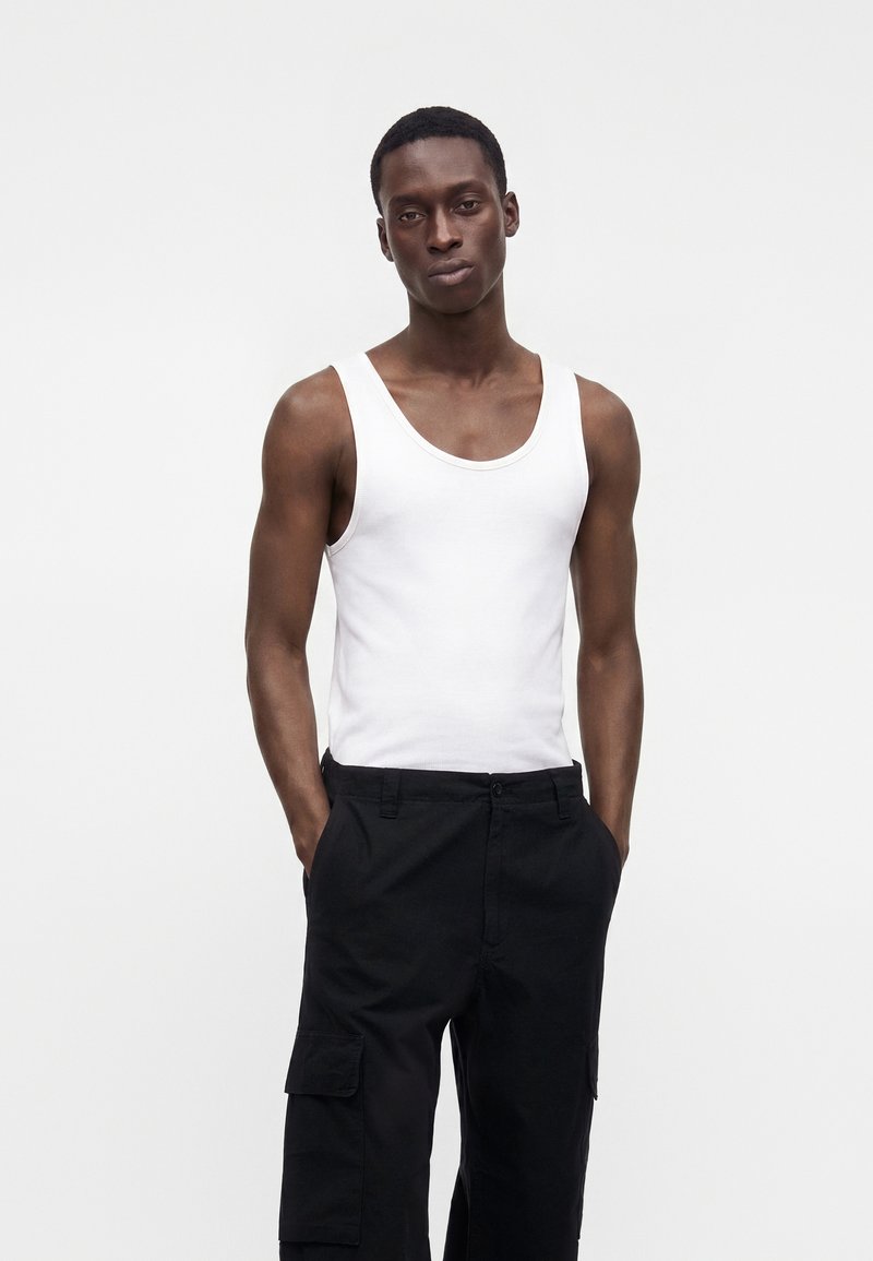 Homme portant un débardeur blanc sans manches et un pantalon cargo noir, les mains dans les poches, sur un fond clair uni.