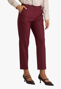 Pantaloni su misura color bordeaux con chiusura a bottone, tasche laterali e design a gamba dritta, abbinati a scarpe con tacco lucide.