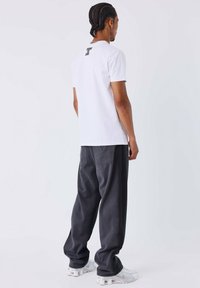 Camiseta blanca con texto en la parte posterior, combinada con pantalones anchos gris oscuro y zapatillas blancas. Conjunto simple y casual con un ajuste relajado.