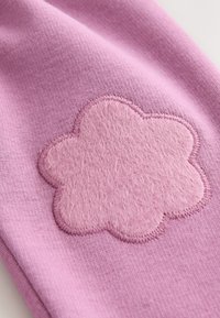 Il tessuto rosa presenta un'applicazione di fiore in feltro morbido, contornata da una cucitura abbinata. La texture complessiva è liscia e leggermente elastica.