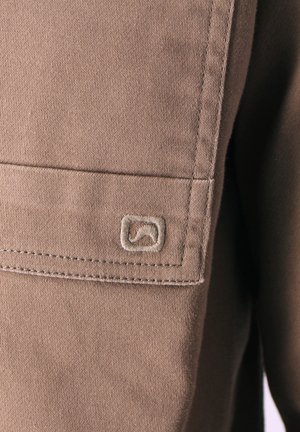 Close-up van een bruine stoffen zak met dubbele stiksels en een klein geborduurd logo in de vorm van een golf bij de hoek.