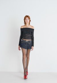 Top negro de encaje fuera de los hombros, mini falda de mezclilla, medias de red y zapatillas de plataforma rojas brillantes con correas en el tobillo. Fondo blanco.