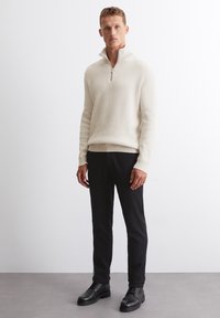 Hellbeige, gerippter Strickpullover mit Kragen und Halbzipp, kombiniert mit schwarzen Hosen und schwarzen Stiefeln, vor einem neutralen Hintergrund stehend.