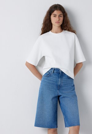 Junge Frau mit langen lockigen Haaren, die ein schlichtes weißes T-Shirt mit kurzen Ärmeln und kniehohe blaue Jeansshorts trägt, steht vor einer weißen Wand.