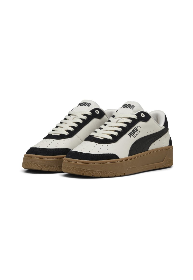 Baskets blanches en cuir avec des accents en suède noir, bout perforé, lacets plats et semelle en caoutchouc brun texturé. Logo PUMA sur les côtés.