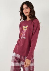 Bordó hosszú ujjú póló egy medve grafikával. Puha anyag, laza szabás. Bordó és fehér kockás pizsamanadrággal párosítva.