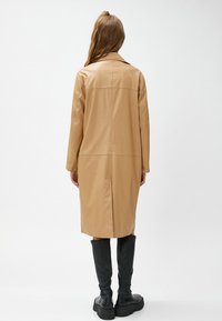 Läderjacka i tan färg med rak design, långa ärmar och sömdetalj på baksidan, i kombination med svarta knäkorta stövlar.