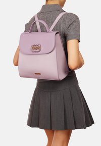 Zaino in pelle lilla con un flap arrotondato, manico superiore e spallacci regolabili. Presenta una chiusura in tono oro e una superficie liscia.