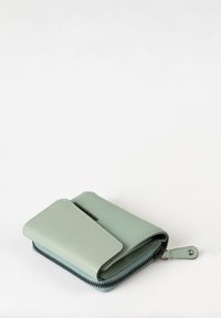 Portafoglio in pelle verde salvia compatto con scomparto con zip, chiusura piegata e un'etichetta con bottone a snap su sfondo bianco.