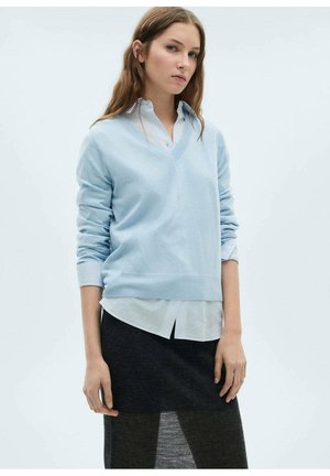Pull en v bleu clair en tissu doux, superposé à une chemise blanche et bleu rayée. La tenue comprend une jupe noire et des manches longues.