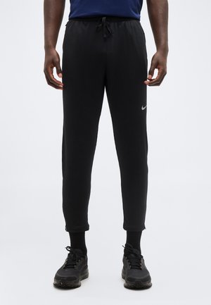 Mees, kes kannab musti Nike fliispükse, millel on pahkluul lukud, musti sokke ja musti tossusid, seisab tavalise heleda tausta ees.