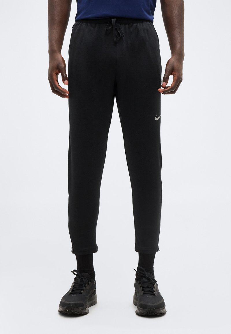 Άνδρας που φοράει μαύρο παντελόνι φόρμας Nike με φερμουάρ στους αστραγάλους, μαύρες κάλτσες και μαύρα αθλητικά παπούτσια, στέκεται μπροστά από ένα απλό ανοιχτό φόντο.