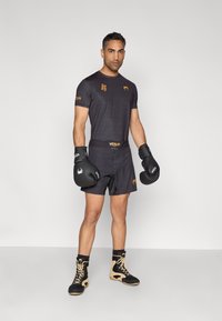 Camisola e calções atléticos pretos, com um padrão subtil; luvas de boxe pretas; botas pretas e bege com atacadores; fundo minimalista.