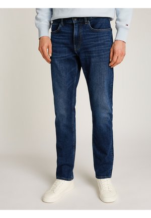 Man die donkerblauwe slim-fit jeans en witte sneakers draagt, staand tegen een effen achtergrond.