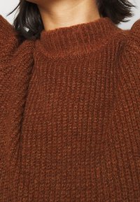 Brun stickad tröja med hög ribbad halsringning och strukturerad tyg, med voluminösa ärmdetaljer och ett varmt, mysigt utseende.