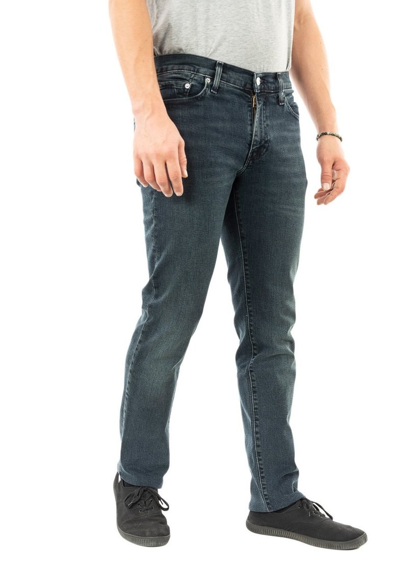 Jeans in denim blu scuro con vestibilità slim, dotati di zip frontale, cinque tasche e dettagli di cucitura sottili. Indossati con sneakers nere.