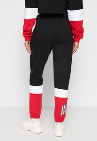 Černé tepláky s červenými a bílými pruhy na manžetách a pase. Mají elastický pas a žebrované detaily. Tisknuté logo na nohavici.