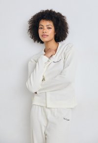 Camisola branca, texturizada, de manga longa com padrão canelado, apresentando um pequeno logótipo bordado. Combinada com calças texturizadas brancas a condizer.