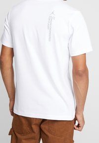 Camiseta de algodón blanca con mangas cortas, cuello redondo y un pequeño logo impreso en gris en la parte superior de la espalda que dice "JORDAN ENGINEERED FOR FLIGHT."