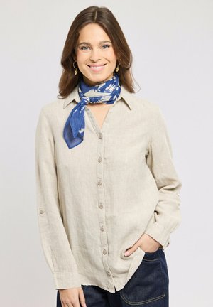 Jeune femme souriante, portant une chemise beige à boutons, une écharpe bleue à motifs nouée autour du cou, et un jean foncé, une main dans la poche.