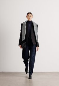 Čierny blazer cez tmavomodrý rolák, šedý šál, široké tmavé nohavice a čierna taška. Elegantné špicaté topánky dopĺňajú celý vzhľad. Jednoduché pozadie.