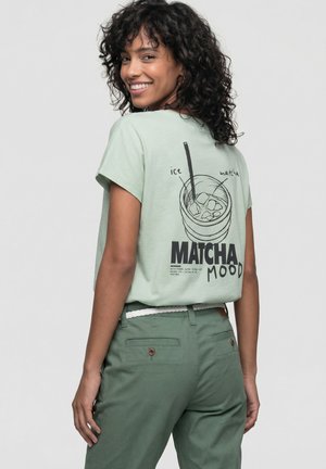Lächelnde Frau mit lockigem Haar, die ein hellgrünes "Matcha Mood"-T-Shirt mit Eistee-Matcha-Grafik trägt, kombiniert mit dunkler grünen Hosen.