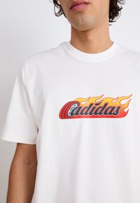 Balts kokvilnas T-kreklis ar lielu sarkanu un oranžu liesmu grafiku, kur zem teksts "Cadidas" ir rakstīts treknā melnā burtiem. Īsas piedurknes, apaļa kakla izgriezums.