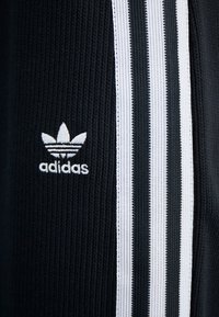 Schwarzer gerippter Stoff mit dem in Weiß gestickten Adidas-Trefoil-Logo neben drei vertikalen weißen Streifen mit schwarzen Akzenten.