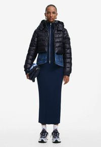 Giubbotto imbottito nero con dettagli in denim, vestito a coste blu navy, calze bianche e sneakers con motivo. Modella che tiene in mano una borsa nera.