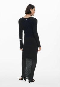 Abito nero in maglia con texture a coste e pannello inferiore trasparente. Presenta maniche lunghe e un collo rotondo, accentuato da un piccolo bottone.