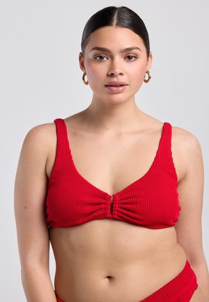 Junge Frau, die ein strukturiertes rotes Bikinioberteil und passende, hoch taillierte Hosen trägt, mit goldenen Creolen an einem einfarbigen hellgrauen Hintergrund.