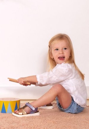 Child wearing a white long-sleeve shirt, denim shorts, and multi-colored sandals, playing with drumsticks beside a yellow patterned drum.