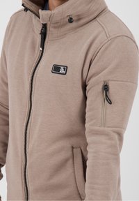 Primo piano di una giacca beige con zip e colletto alto, piccola toppa nera con la scritta "WE ARE ALIFE" e tasca con zip sulla manica superiore.