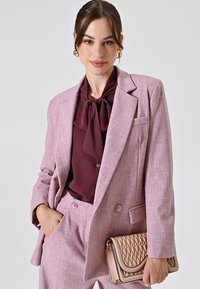 Hellrosa maßgeschneiderter Blazer mit zwei Knöpfen, kombiniert mit einer burgunderfarbenen Bluse mit einer Schleife am Ausschnitt. Hält eine gesteppte beigen Handtasche.