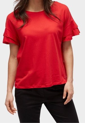 Vrouw die een rood topje met korte mouwen en gelaagde ruches draagt, en een zwarte broek. Afgebeeld van nek tot heupen tegen een grijze achtergrond.