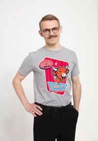 Grå bomulds t-shirt med et farverigt grafik af en ko og teksten "La Vache qui Rit" i forskellige skrifttyper, iført med sorte bukser.