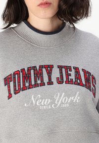 Felpa grigia con colletto a costine. Presenta il testo a quadri rosso e nero "TOMMY JEANS" e ricamo bianco "New York dal 1985". Texture morbida.