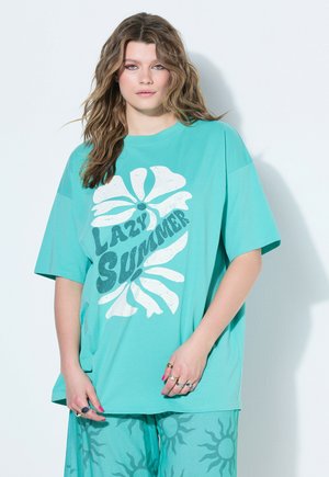 Junge Frau mit welligem Haar trägt ein übergroßes T-Shirt in Türkis mit dem Schriftzug „Lazy Summer“ und floralem Design, kombiniert mit türkisen Hosen mit Sonnenmustern.